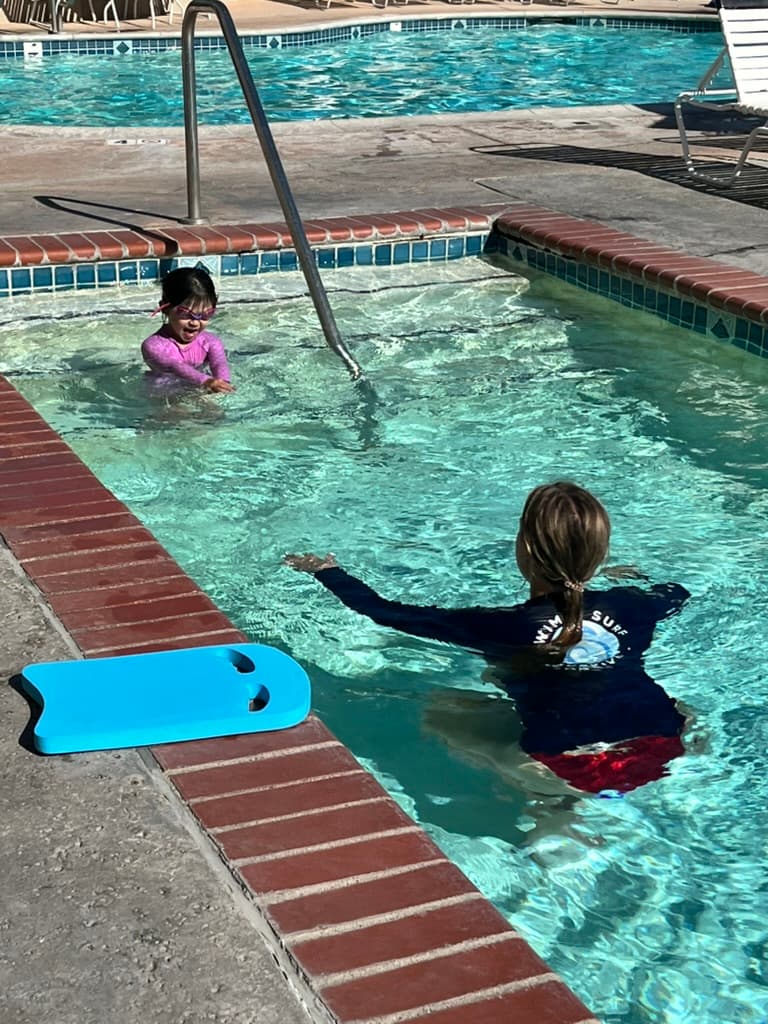 Swim instructor guiding a young child with a kickboard in a sunlit pool
